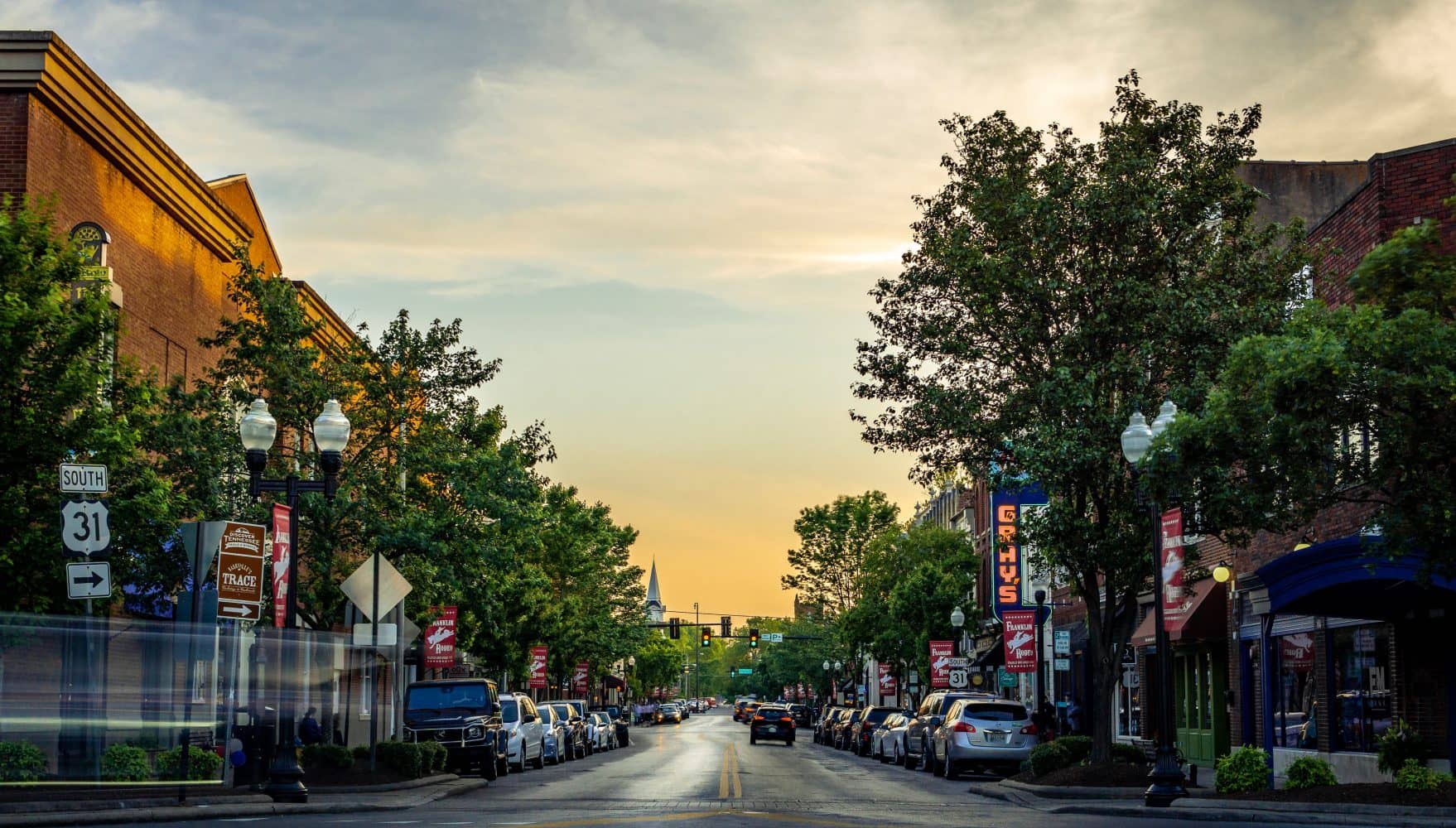 View of city street with cars, trees, buildings, sky, and church steeple. View of city street with cars, trees, buildings, sky, and church steeple.