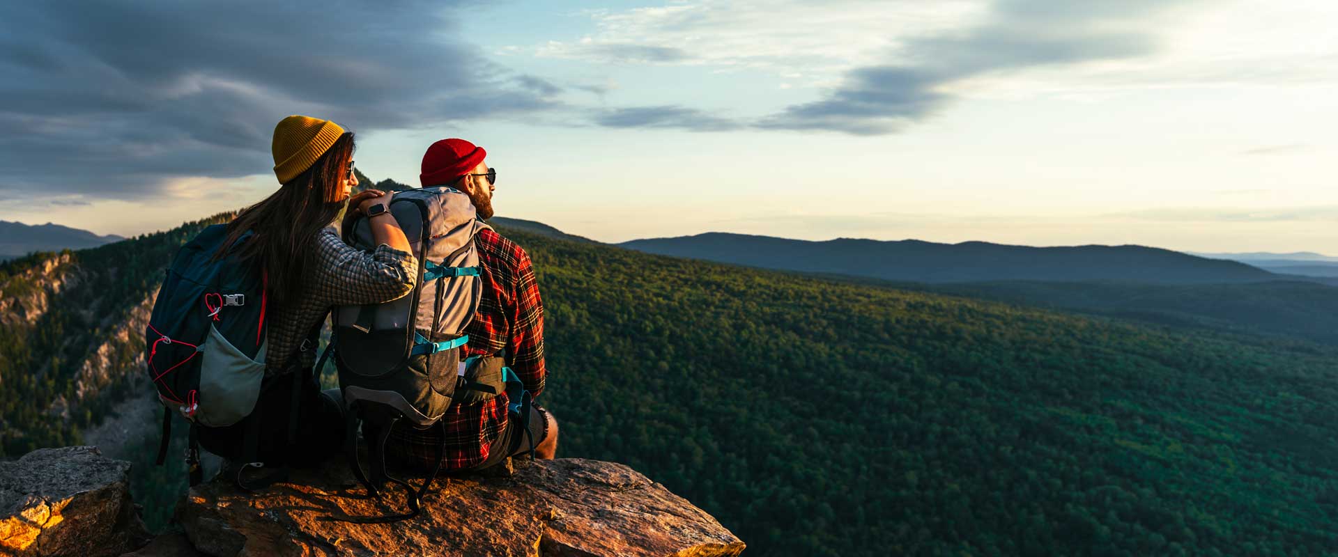 Two travel nurses wearing backpacks sitting on the mountain top overlooking the sky.