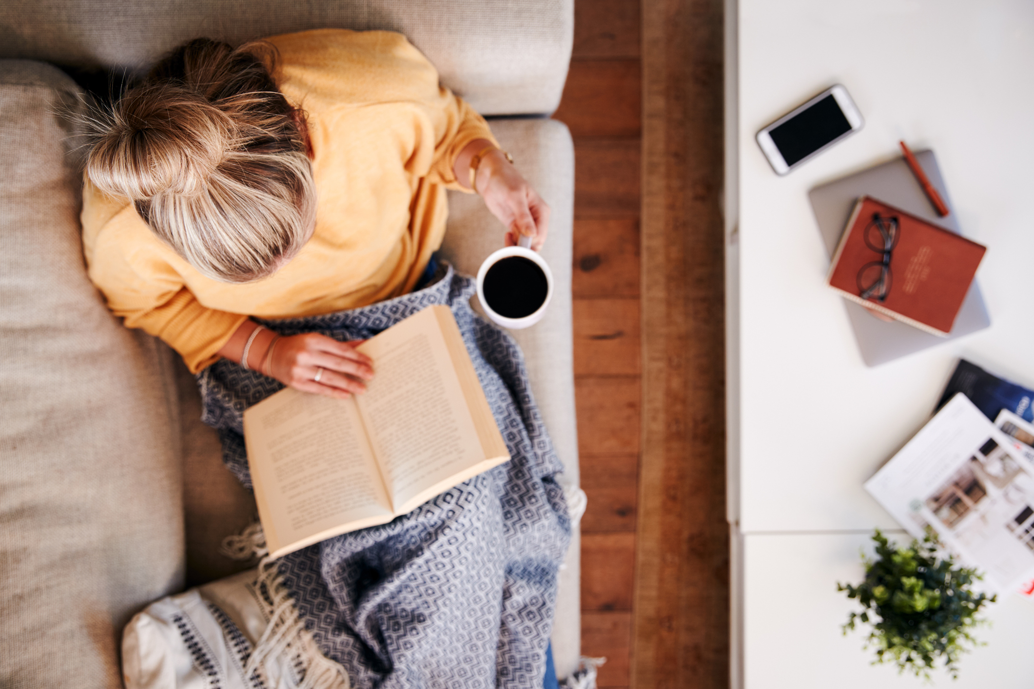 woman curls up under blanket on sofa with book and hot tea woman curls up under blanket on sofa with book and hot tea