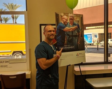 TOM Stephen Pietzak PT Month at work, smiling next to poster of him with patient 