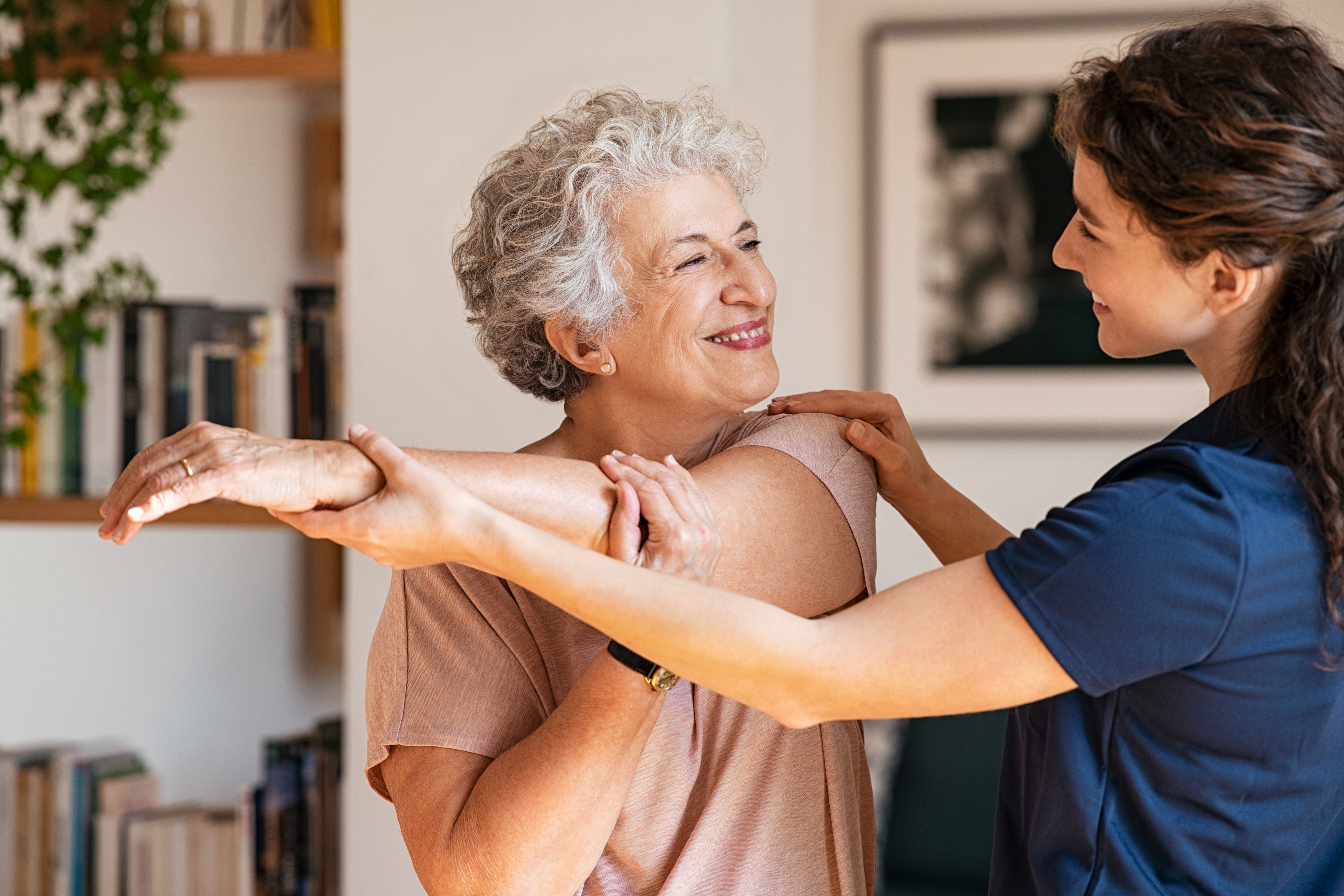 Happy female nurse wearing blue scrubs helping her elderly, female patient with a stretching exercise.